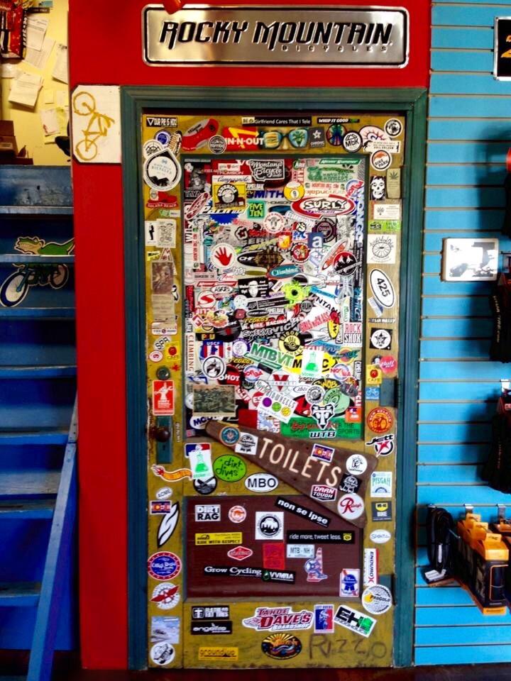 A brightly colored door covered in an assortment of stickers from various brands and organizations, with the words "TOILETS" prominently displayed on a wooden sign. The door is framed by a teal border, and the background features a blue wall with bike accessories hanging nearby. The top of the door displays a metal sign reading "ROCKY MOUNTAIN."