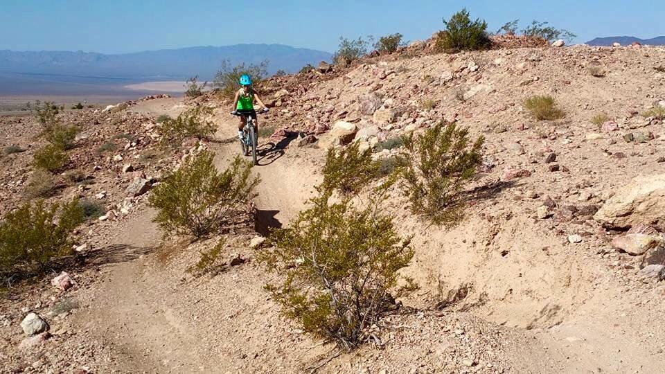 A mountain biker riding along a dirt trail in a rocky landscape, surrounded by desert vegetation and distant mountain ranges under a clear blue sky. Bootleg Canyon mountain bike trail.