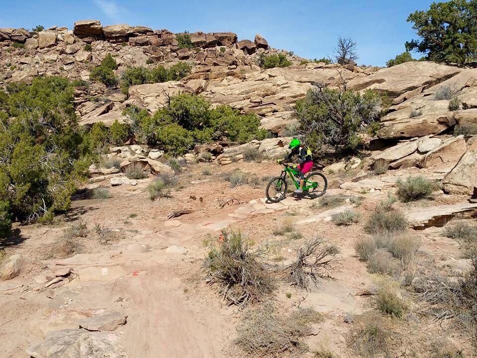 A person riding a green mountain bike along a rocky trail surrounded by sparse vegetation and large boulders under a clear sky. Klondike Bluffs mountain bike trail.
