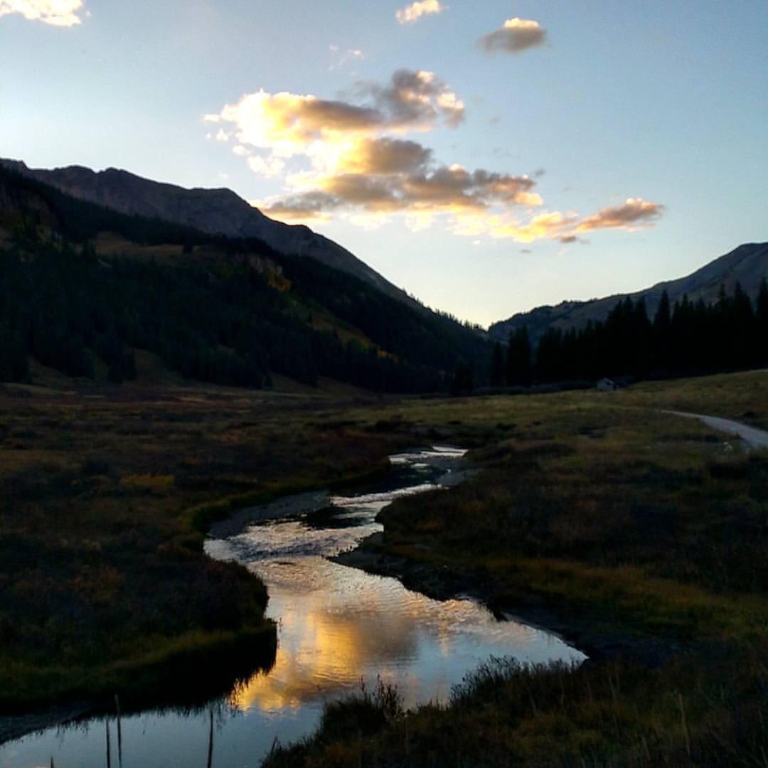 A serene landscape featuring a meandering stream reflecting clouds and mountains at twilight. Surrounding the stream are lush meadows and a backdrop of forested hills, with the sky transitioning from daylight to evening hues. Trail 401 mountain bike trail.