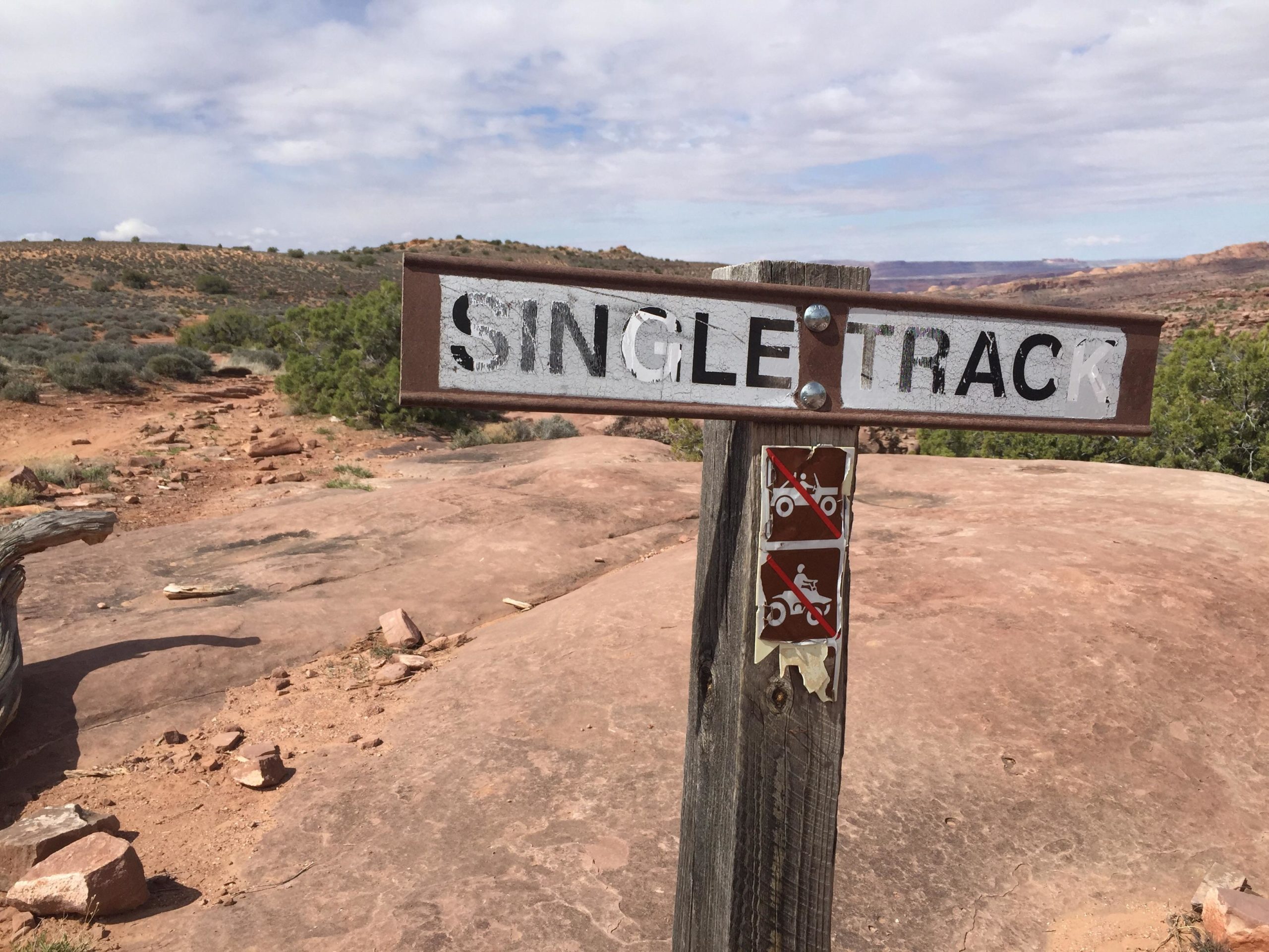 A weathered wooden sign marking a single track trail in a rocky, arid landscape, with sparse vegetation and distant hills under a cloudy sky. The sign features the words "SINGLE TRACK" in bold letters and has no motor vehicle symbols below it, indicating prohibited areas for ATVs and motorcycles. Porcupine Rim mountain bike trail.