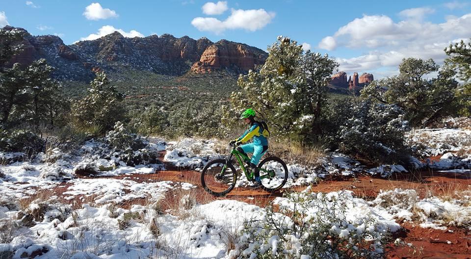 A person riding a mountain bike along a snowy, red dirt trail, surrounded by evergreen trees and rocky mountains in the background. The sky is partly cloudy, and the scene captures the beauty of a winter landscape. Bell Rock Trailway mountain bike trail.
