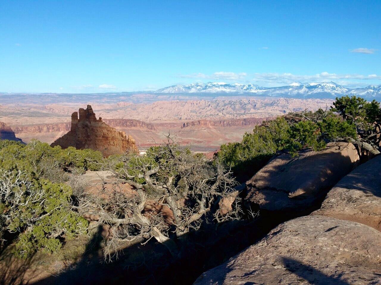 A panoramic view of a rugged landscape featuring a prominent rock formation in the foreground, surrounded by sparse vegetation. In the background, expansive canyons and mountains are visible under a clear blue sky dotted with a few clouds. The scene captures the natural beauty and vastness of the terrain. Dead Horse Point State Park mountain bike trail.