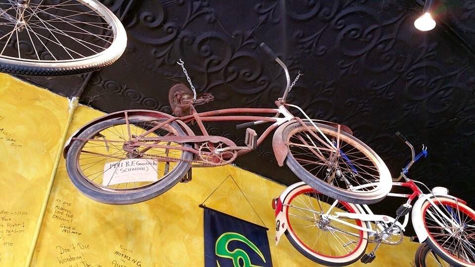 A vintage bicycle hanging from the ceiling against a yellow wall, featuring a sign that reads "1941 B.F. Goodrich (Schwinn)." Other bicycles can be seen in the background, showcasing their colorful designs. The ceiling has an ornate black texture.