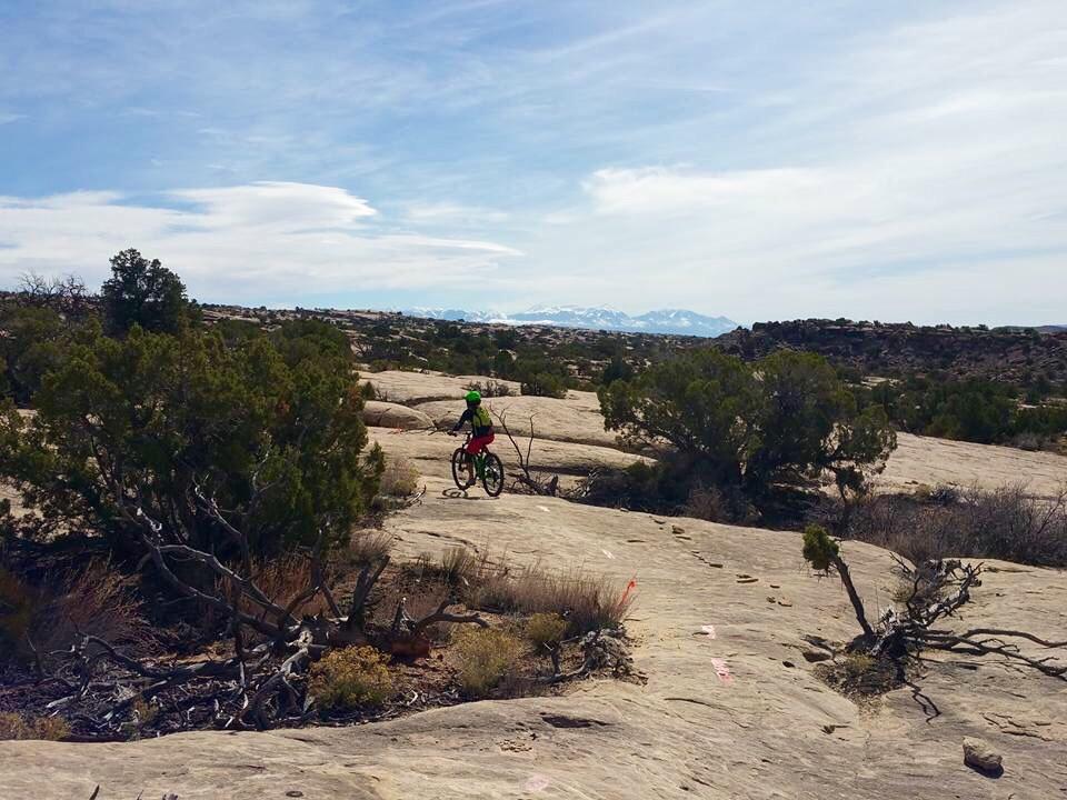 A mountain biker rides on rocky terrain surrounded by shrubs and distant mountains under a partly cloudy sky. Klondike Bluffs mountain bike trail.