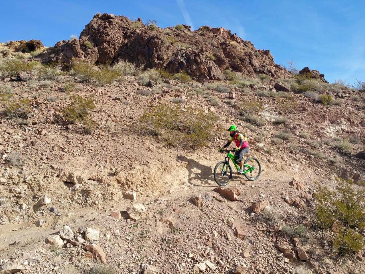 A person riding a green mountain bike along a rocky trail in a desert landscape, with rugged hills and sparse vegetation in the background under a clear blue sky. Bootleg Canyon mountain bike trail.