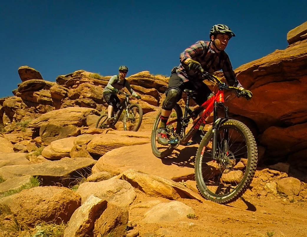 Two mountain bikers navigating a rocky trail in a desert landscape. One biker is jumping over a rock, while the other is riding on a nearby path. The sky is clear and blue, and the terrain consists of orange-red rocks and sparse vegetation. Captain Ahab mountain bike trail.