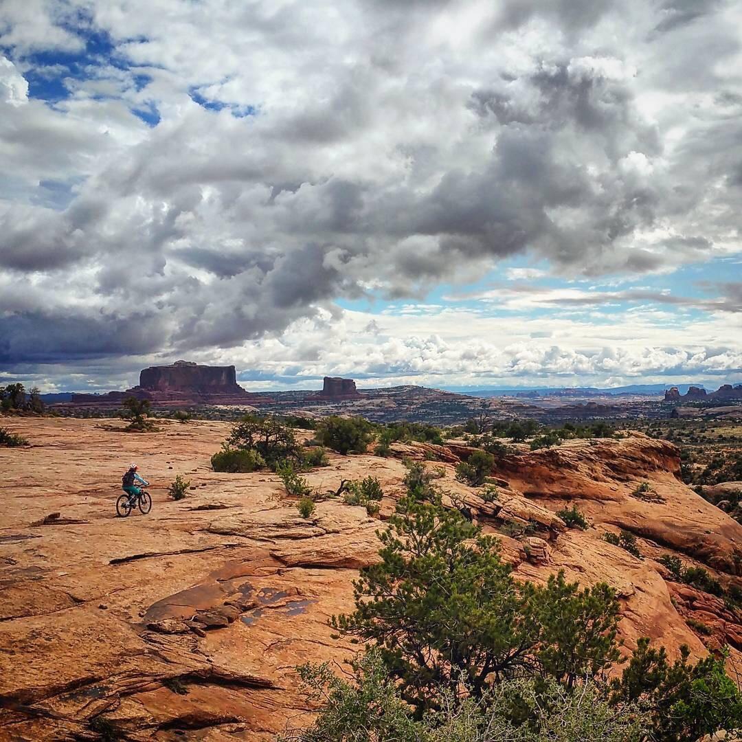 A cyclist rides on rocky terrain with expansive desert scenery in the background, featuring large mesas and a cloudy sky. Green vegetation peeks through the rugged landscape, showcasing a blend of nature and outdoor adventure. Navajo Rocks mountain bike trail.