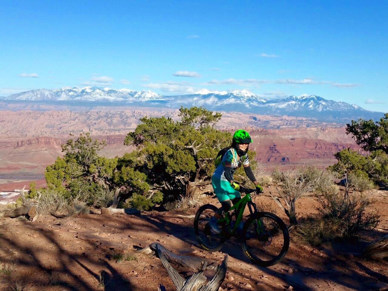 A mountain biker in a green helmet and outfit rides along a rocky trail, surrounded by desert vegetation and stunning mountain scenery in the background. Snow-capped peaks rise in the distance under a bright blue sky. Dead Horse Point State Park mountain bike trail.
