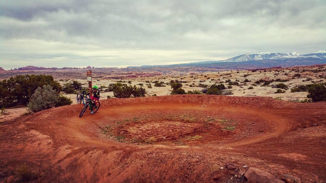A cyclist riding on a dirt biking trail with a circular turn in a rugged, desert landscape. The scene features rocky terrain, sparse vegetation, and a cloudy sky with distant mountains. A trail sign is visible nearby. Moab Brand Trails mountain bike trail.