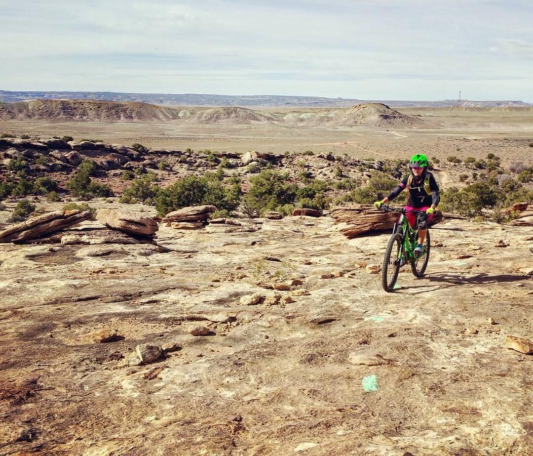 A person riding a mountain bike on rocky terrain, surrounded by a vast landscape of hills and sparse vegetation under a partly cloudy sky. The rider is dressed in black and red cycling gear, wearing a green helmet, and appears to be navigating the rugged ground. Klondike Bluffs mountain bike trail.