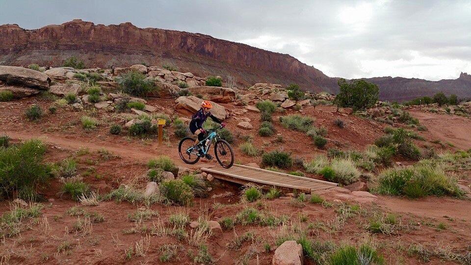 A mountain biker in an orange helmet rides over a wooden bridge on a dirt trail surrounded by rocky terrain and sparse vegetation, with rugged cliffs visible in the background under a cloudy sky. Moab Brand Trails mountain bike trail.