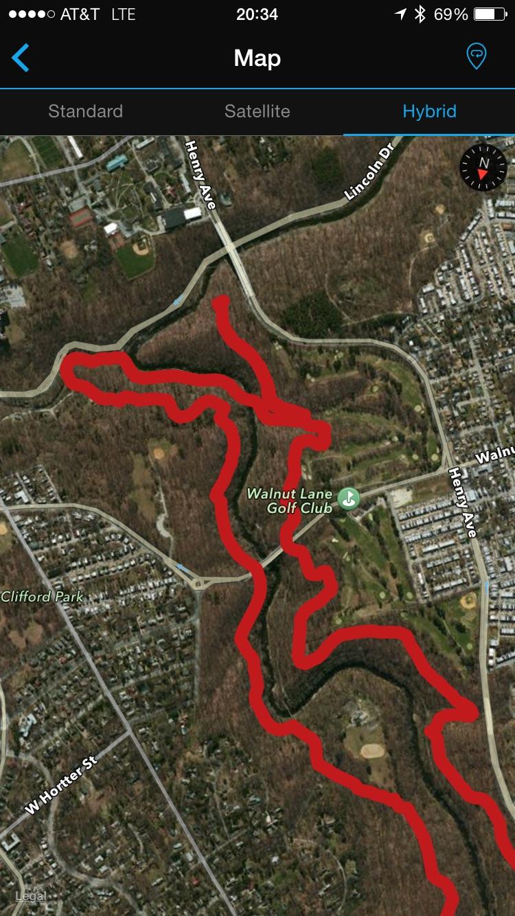 Hybrid map view showing a winding river path outlined in red, surrounded by wooded areas and residential neighborhoods. Key landmarks include the Walnut Lane Golf Club, athletic fields, and various streets such as Henry Avenue and Lincoln Drive. The image captures a blend of natural and urban environments. Wissahickon Valley Park mountain bike trail.