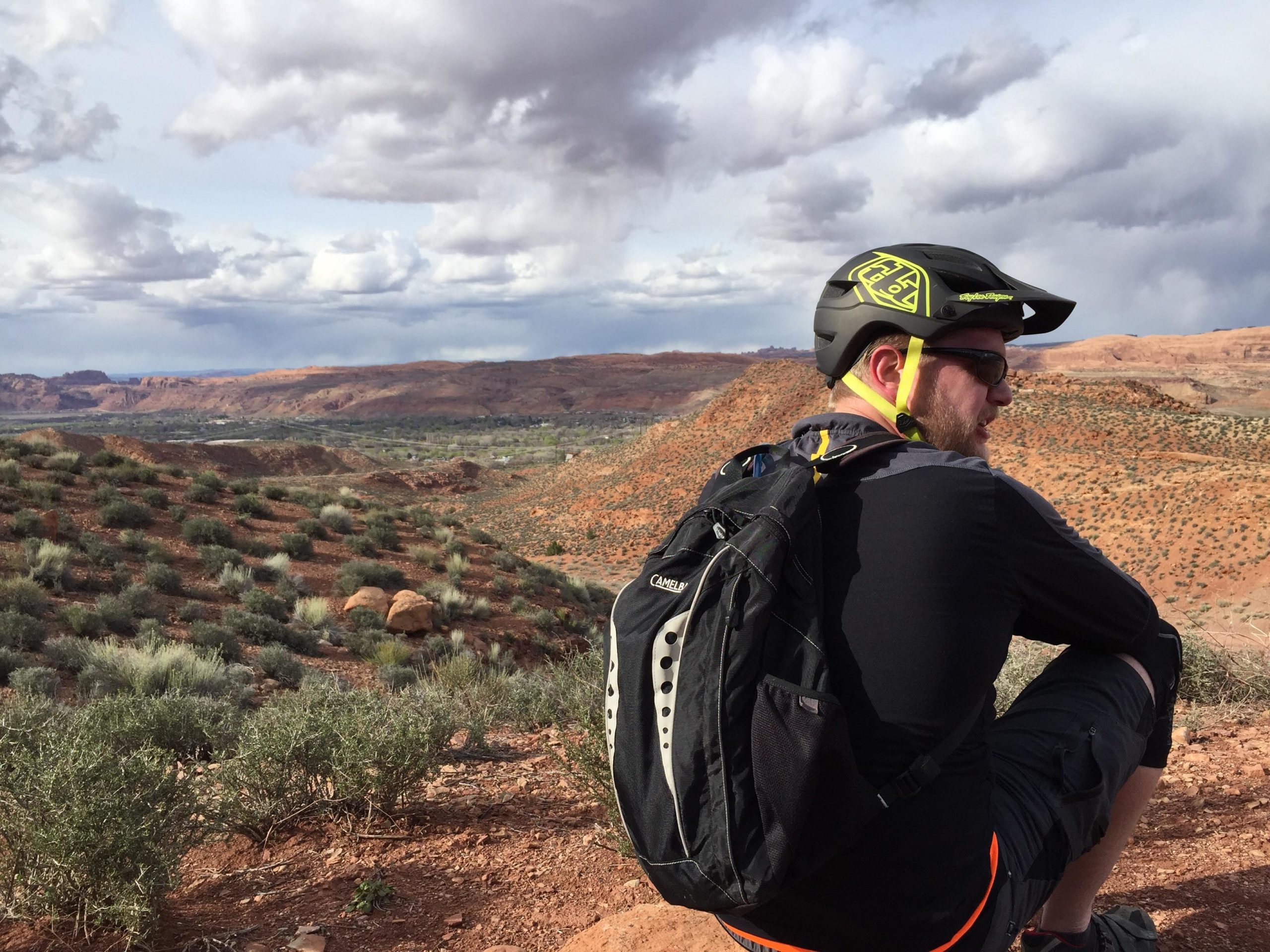 A person wearing a helmet and sunglasses sits on a rocky outcrop, gazing out over a sprawling desert landscape dotted with shrubs and hills under a cloudy sky. Pipe Dream mountain bike trail.