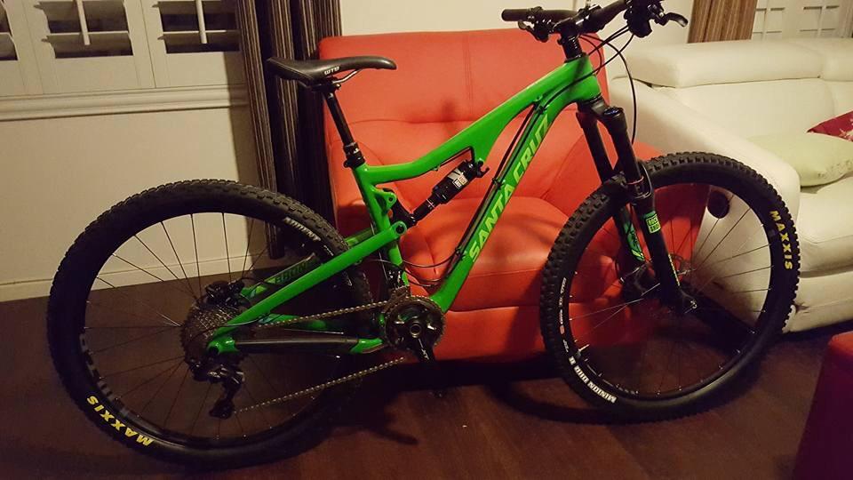 Santa Cruz Bronson: A vibrant green mountain bike leaning against a red couch, with a sleek design featuring thick tires and a suspension system. The bike's frame is prominently labeled "Santa Cruz." The background includes white walls and a second couch, contributing to a cozy indoor setting.