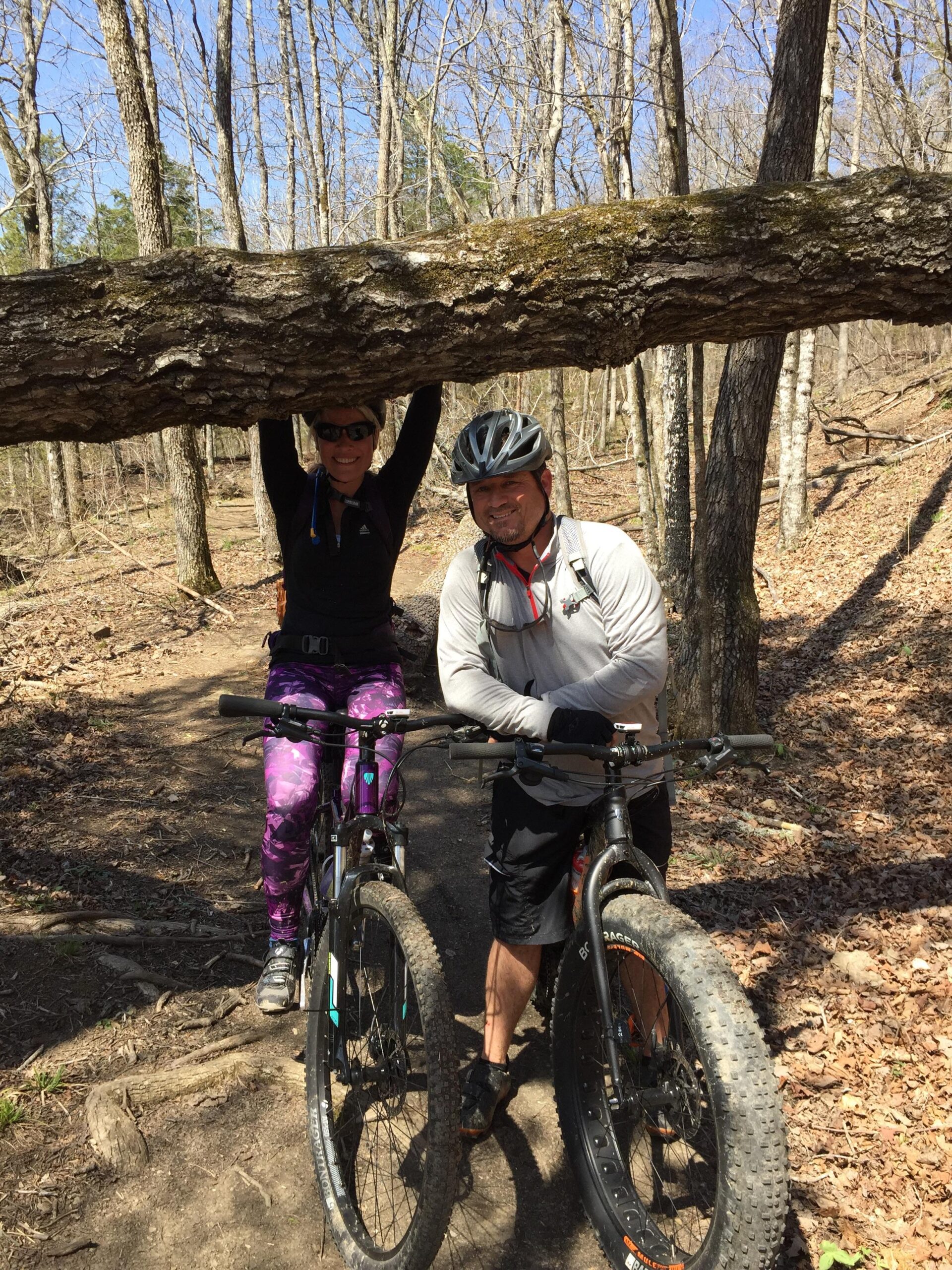 Two mountain bikers posing for a photo on a wooded trail. A fallen tree log is balanced above the woman, who is playfully hanging from it while wearing sunglasses and colorful leggings. The man, wearing a helmet and gloves, stands next to her, smiling. Both bikes are parked beside them, surrounded by leafy ground and trees under a clear blue sky. Fossil Flats mountain bike trail.