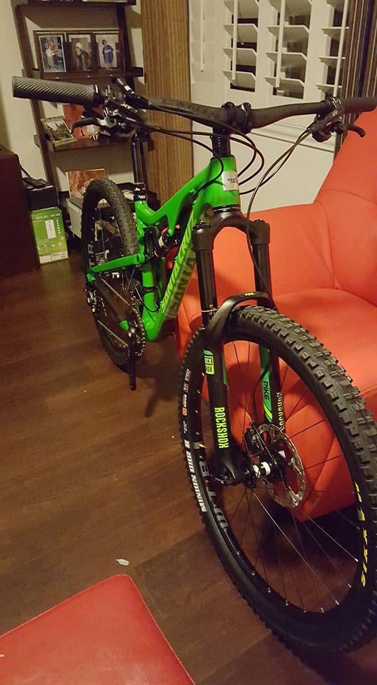 Santa Cruz Bronson: A bright green mountain bike is positioned in a living room, leaning against a red couch. The bike features thick tires with a detailed tread pattern, dual suspension, and disc brakes. In the background, there are shelves displaying framed photos and other decor, with warm wooden flooring visible.