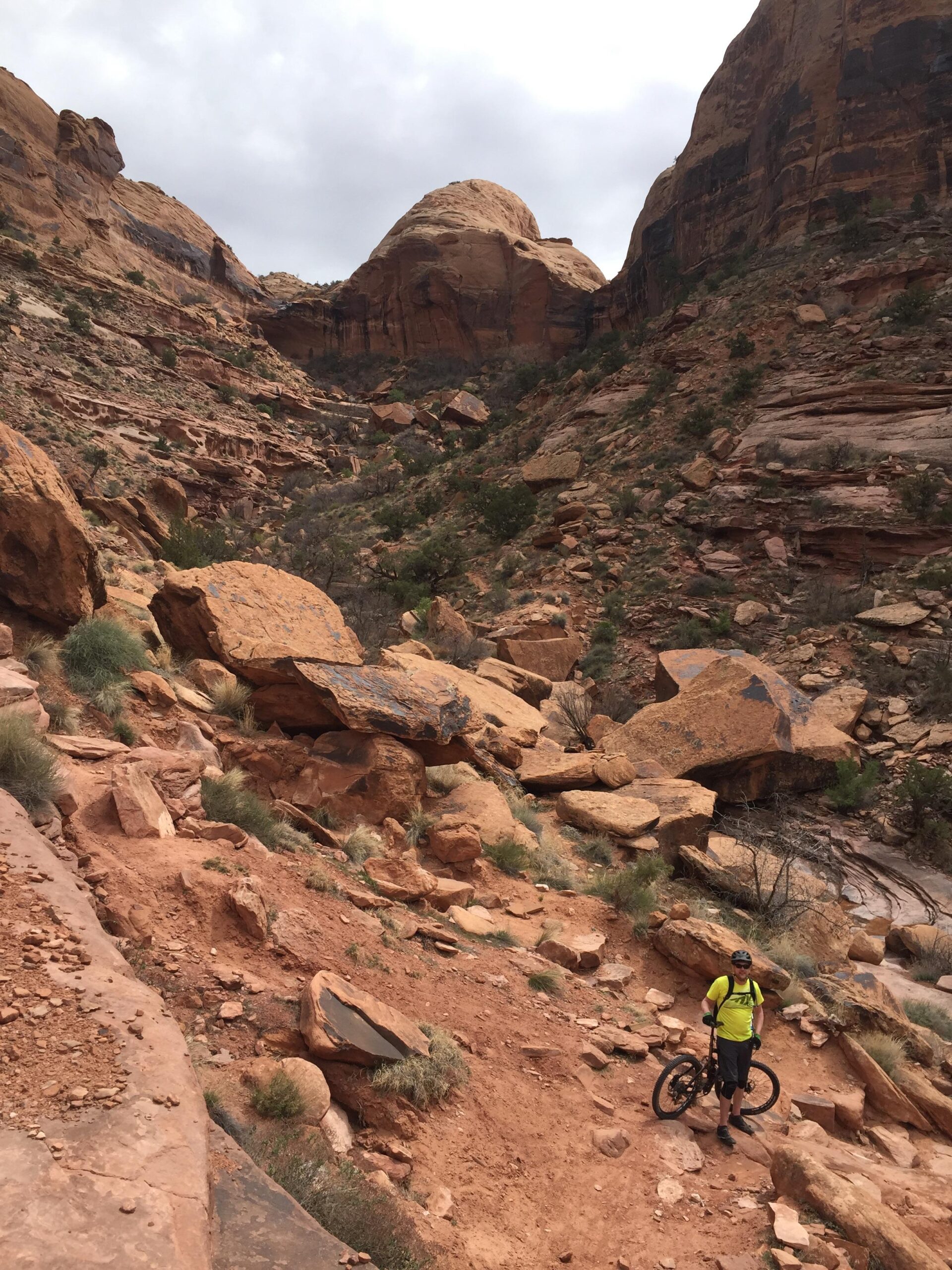 A person in a yellow shirt stands next to a mountain bike on a rocky trail surrounded by red sandstone formations and sparse vegetation. The scene is set against a backdrop of steep canyon walls under a cloudy sky. Porcupine Rim mountain bike trail.