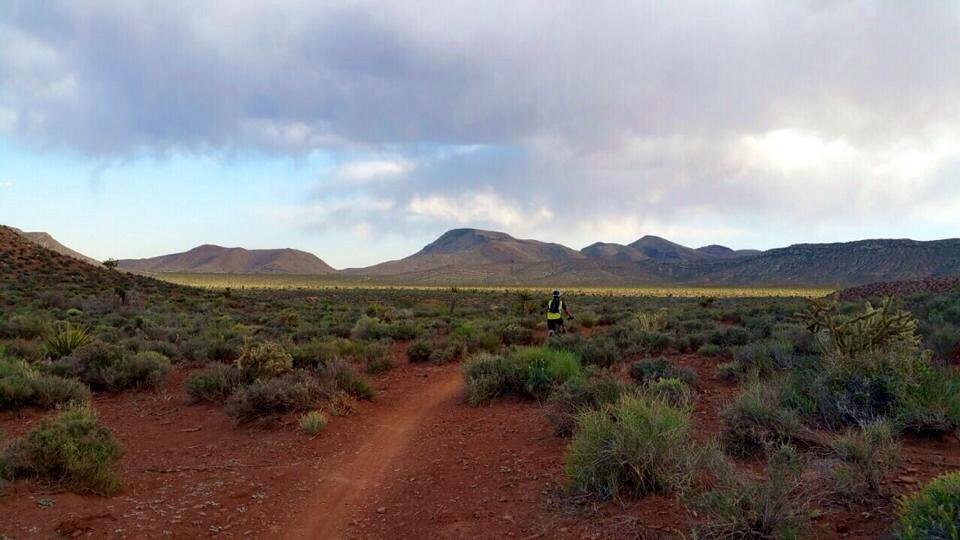 A person walking along a dirt path in a vast landscape featuring low shrubs and red soil, with mountains visible in the distance under a partly cloudy sky. Blue Diamond mountain bike trail.