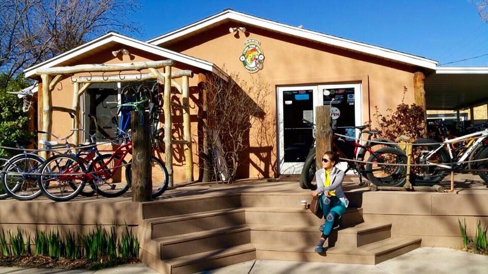 A woman sits on the steps of a bike shop, enjoying a coffee. The shop features a rustic design with bicycles parked outside and a wooden structure for securing bikes. The exterior is painted in warm earthy tones, surrounded by greenery and a clear blue sky.