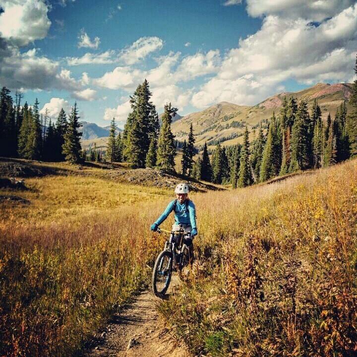 A person in a blue long-sleeve shirt and helmet is riding a mountain bike along a dirt trail in a vibrant, grassy landscape with trees and mountains in the background under a partly cloudy sky. Trail 401 mountain bike trail.