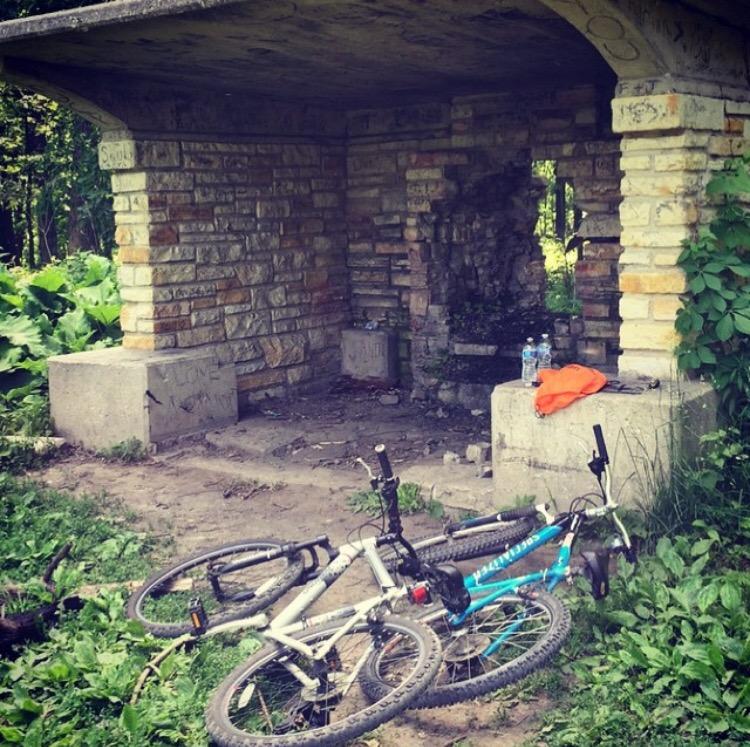 Specialized Rockhopper: An abandoned stone shelter surrounded by lush greenery, with two bicycles lying on the ground in front. The shelter has a partially collapsed wall and contains some debris, while a few water bottles and an orange item are placed on a concrete surface inside.