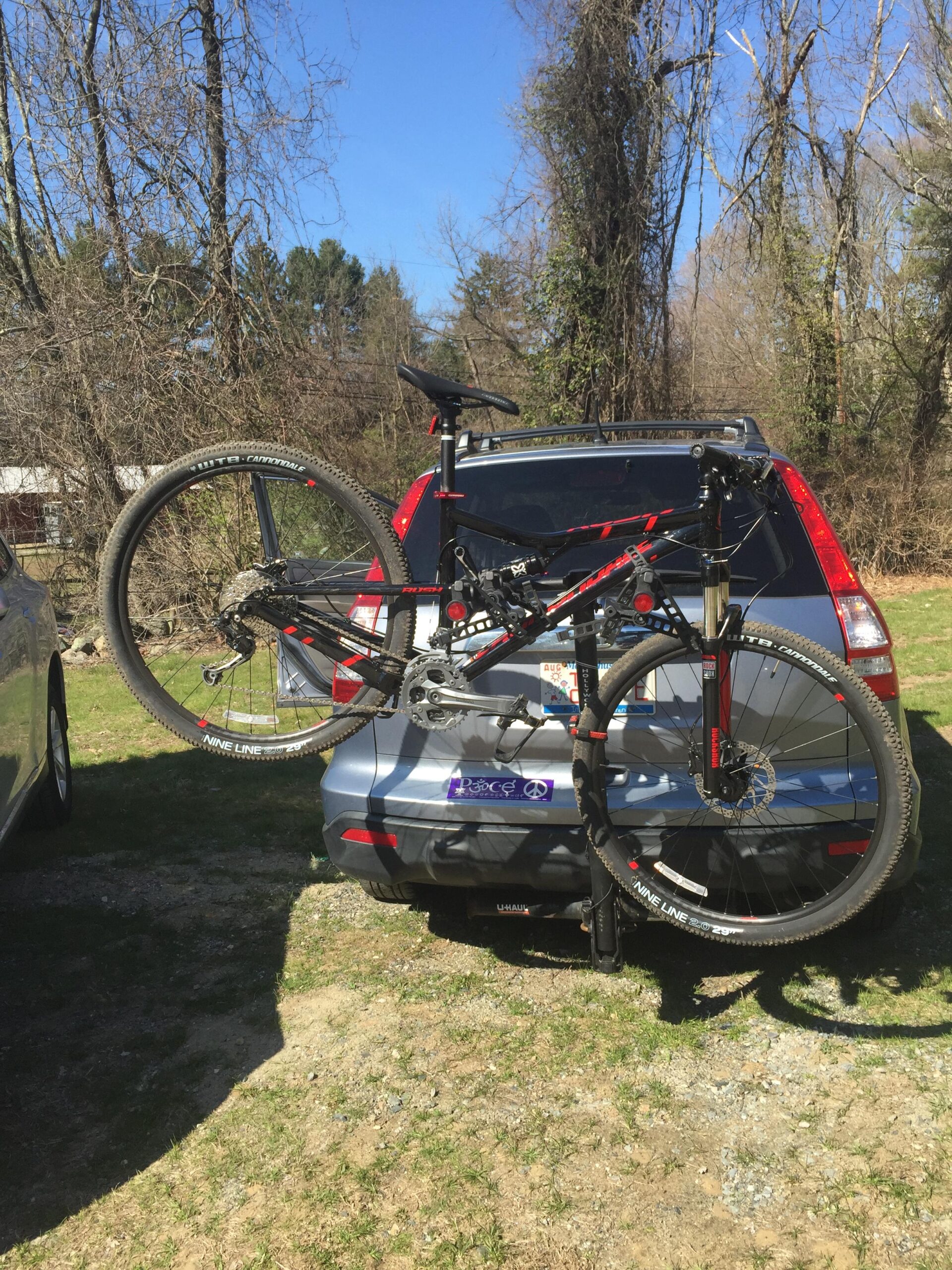 Cannondale Rush 29 1: A mountain bike is mounted on the back of a blue SUV parked on a gravel surface. The bike is positioned upside down with the front wheel in the air. In the background, there are trees with no leaves, indicating early spring or late winter. The sky is clear and blue, adding a bright atmosphere to the scene.