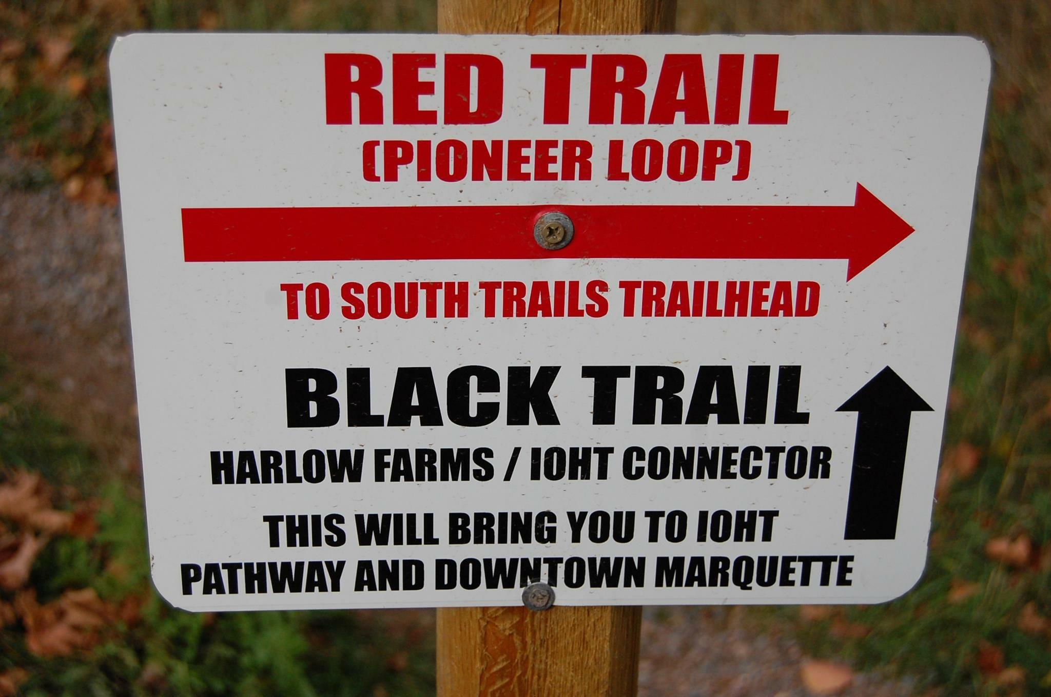 Signpost indicating trail directions, featuring labels for the Red Trail (Pioneer Loop) leading to the South Trails trailhead and the Black Trail connecting to Harlow Farms and the IOHT, with arrows indicating the respective directions. Noquemanon Trails Network: South Marquette Trails mountain bike trail.