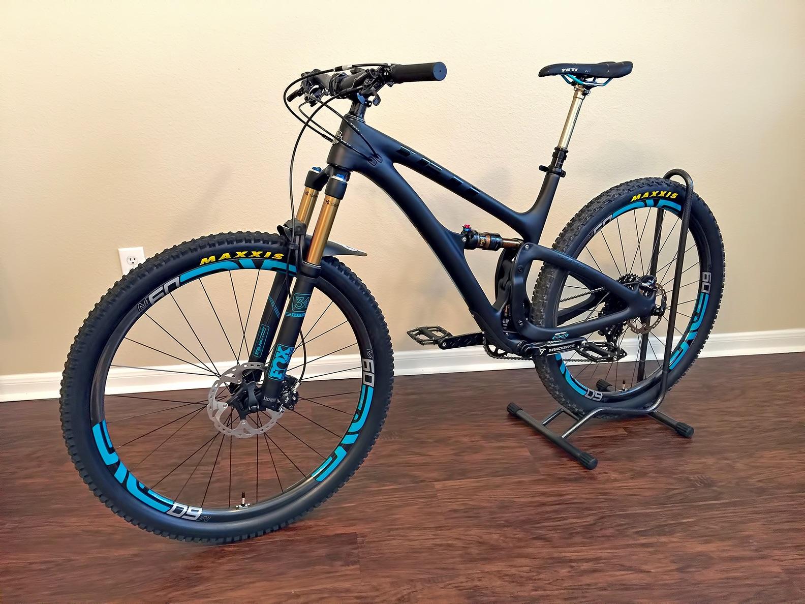 Yeti SB4.5c: A sleek mountain bike with a black and blue frame, equipped with front suspension forks and prominent Maxxis tires. The bike is positioned on a stand, showcasing its features against a neutral wall and wooden floor. The bike components include disc brakes and a high-performance saddle.