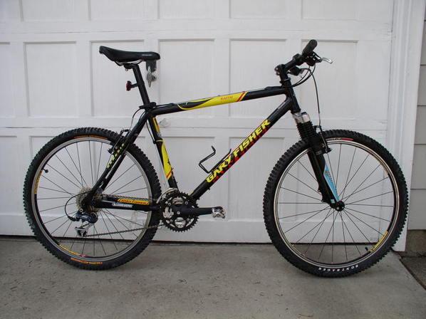 Gary Fisher Kai Tai: Mountain bike with a black frame featuring yellow and red accents, positioned against a white garage door. The bike has knobby tires, front suspension, and a single gear setup, suggesting it's built for off-road riding.