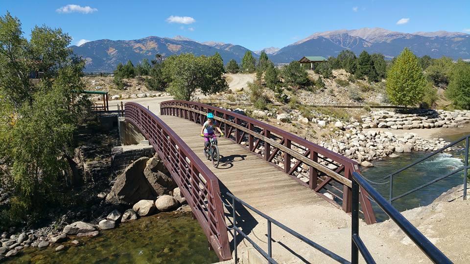 A cyclist rides across a wooden bridge surrounded by trees and a river, with mountains in the background under a clear blue sky. Midland Hills Trails mountain bike trail.