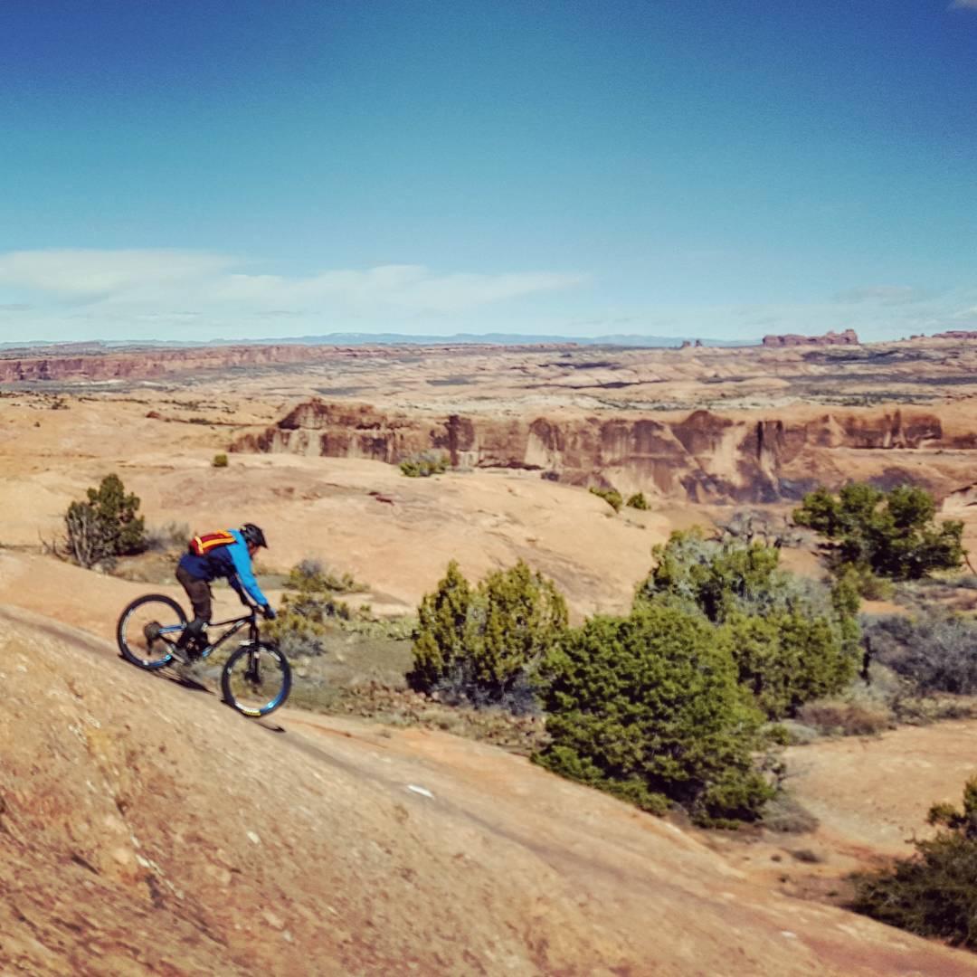 A mountain biker dressed in a blue jacket and helmet rides along a rocky trail overlooking a vast, arid landscape with distant cliffs and sparse greenery under a clear blue sky. Slickrock mountain bike trail.