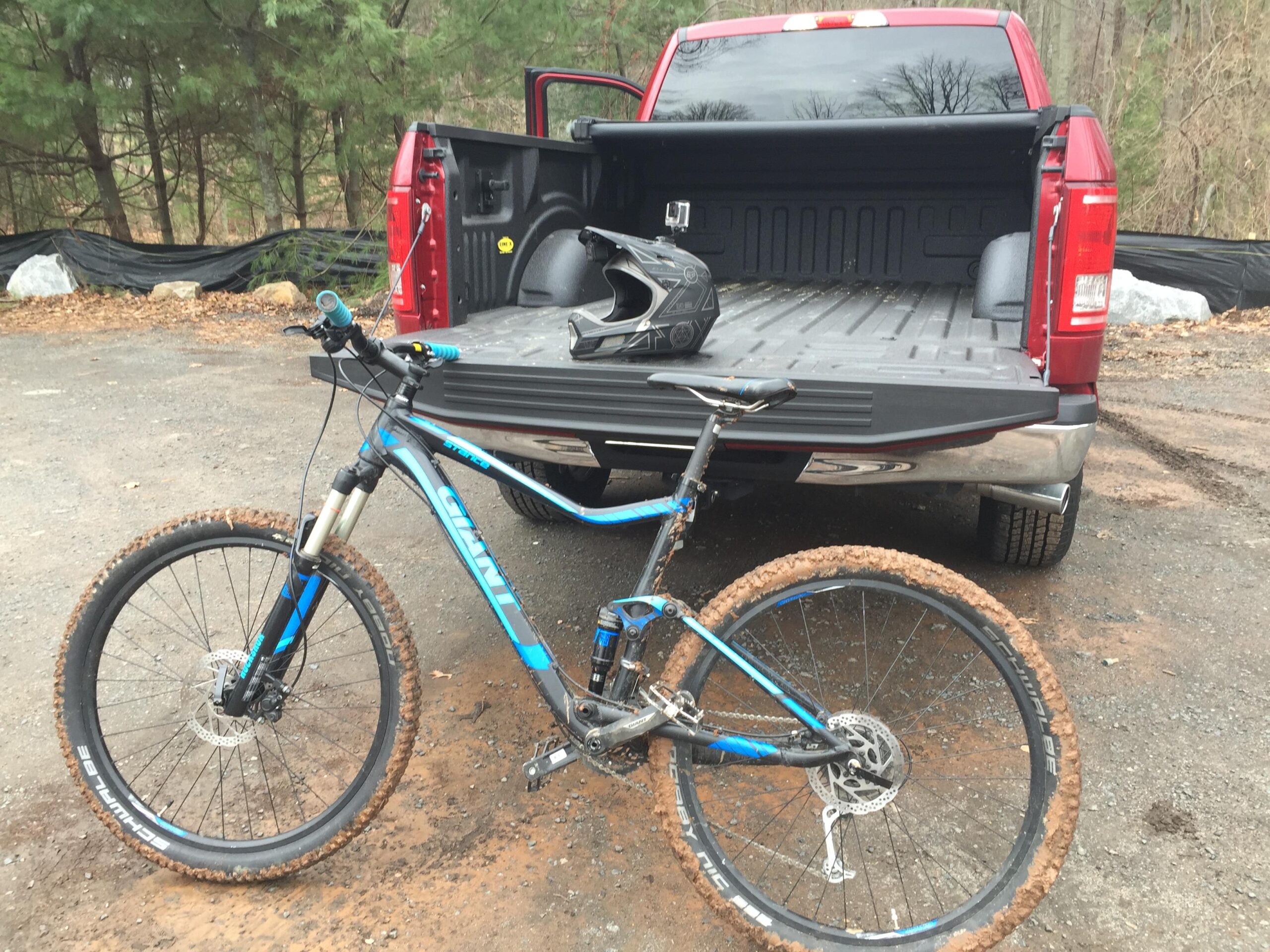 Giant Stance 27.5 2: Alt tag description: A mountain bike with mud on its tires is leaning against the tailgate of a red pickup truck. A black helmet is placed on the truck bed, and the background features trees and a gravel area.