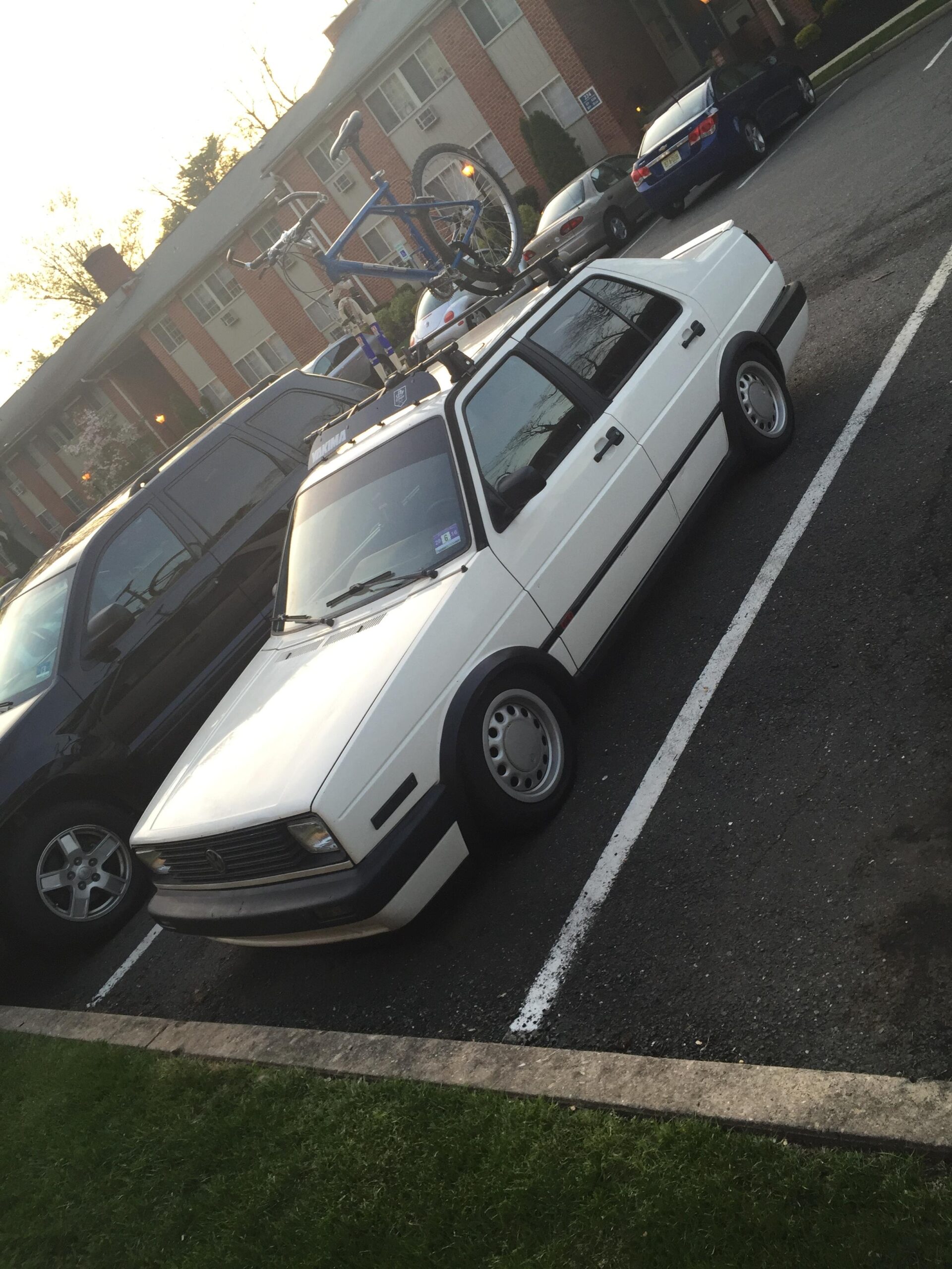 GT Ricochet: A white car parked in a lot with a blue bicycle secured on the roof rack. The background includes brick apartment buildings and other parked vehicles. The setting appears to be during the evening with soft lighting.