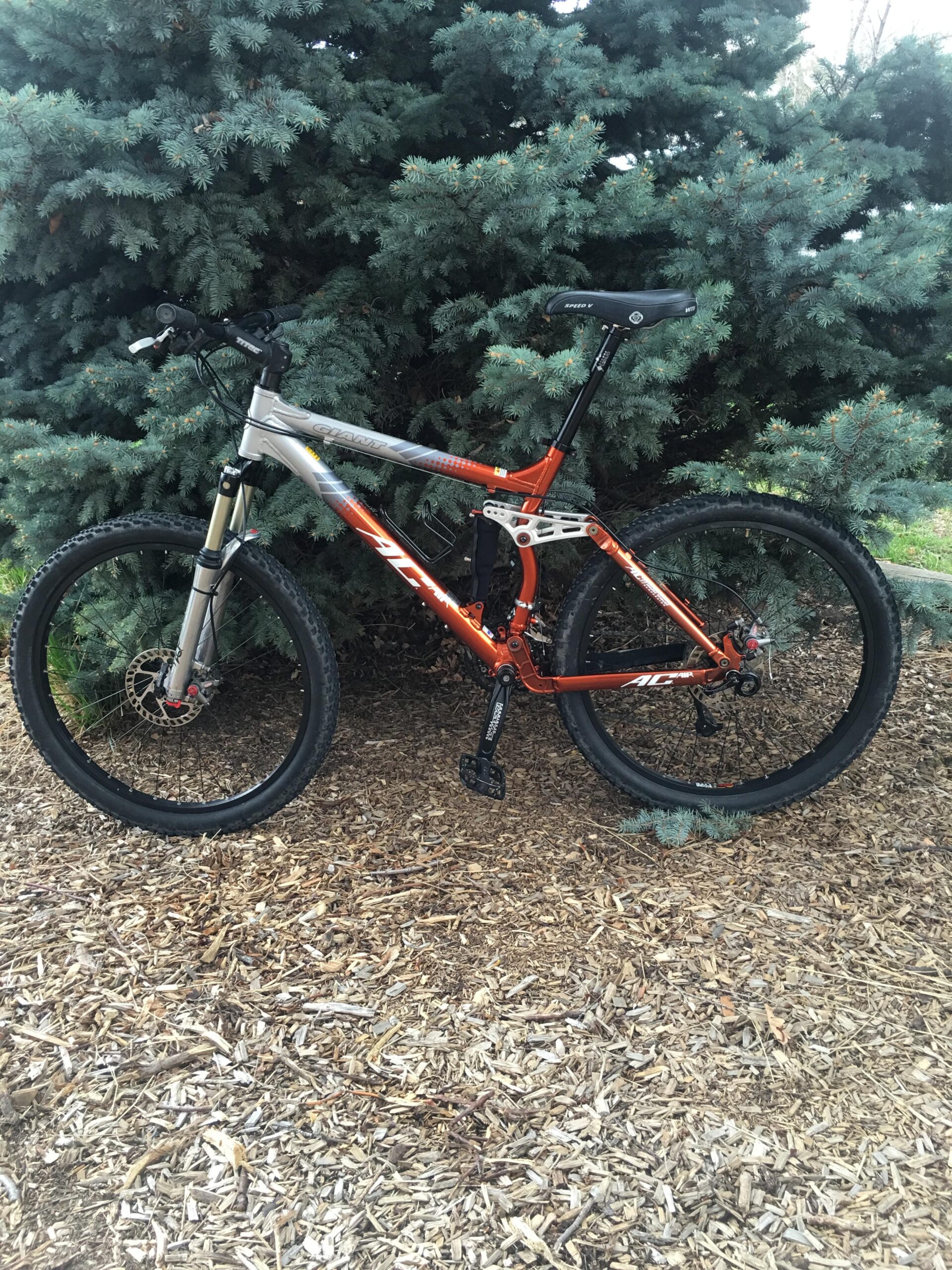 Giant AC: A mountain bike with a silver and orange frame stands on a bed of mulch, surrounded by evergreen trees. The bike features front suspension and thick black tires, emphasizing its rugged design suitable for off-road use.
