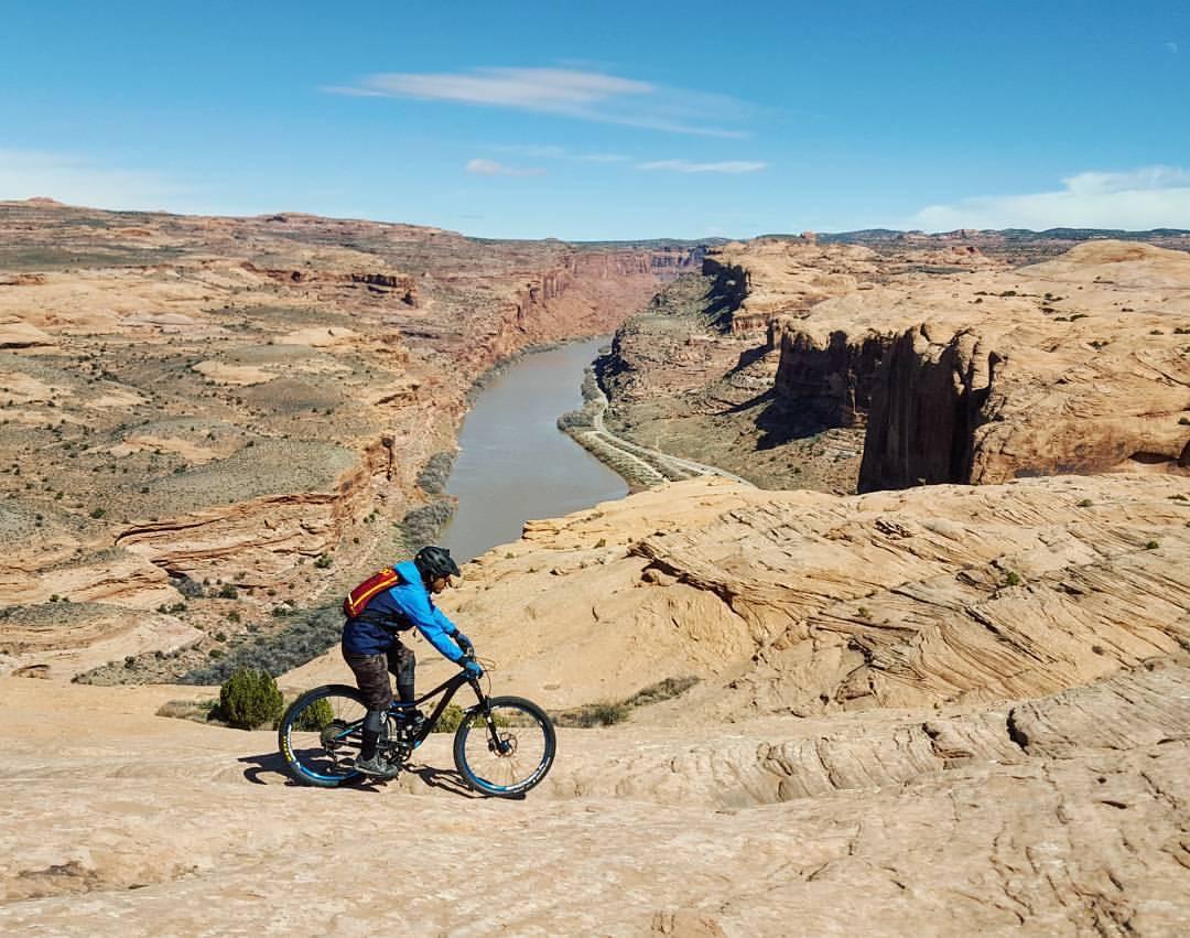 A mountain biker rides along a rocky terrain overlooking a river canyon, with steep cliffs and a clear blue sky in the background. Slickrock mountain bike trail.