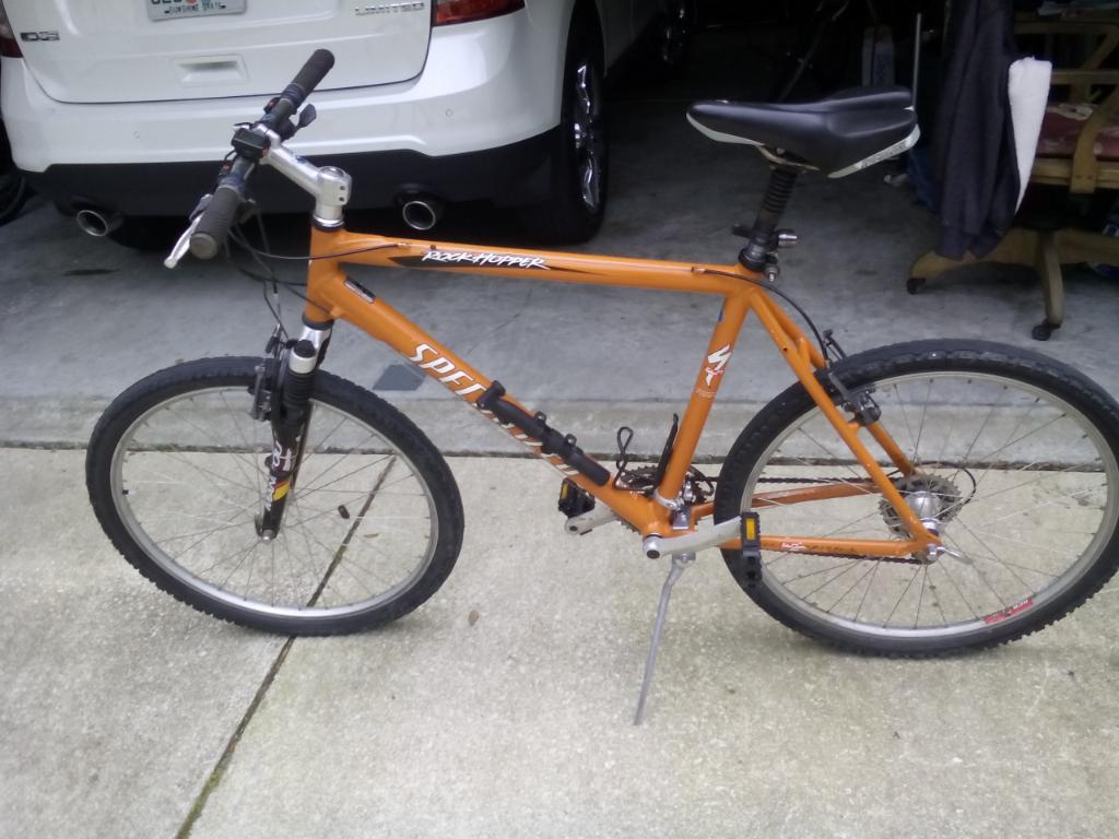 Specialized Rockhopper: A mountain bike with an orange frame and black wheels is parked on a concrete surface. The bike features a front suspension, black handlebars, and a comfortable seat. In the background, a white vehicle is partially visible, along with some outdoor furniture.