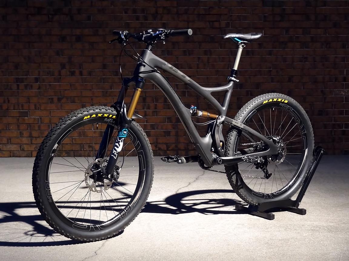 Yeti SB5c: Alt text: A modern mountain bike with a sleek design, featuring a black and gray frame, front suspension fork, and Maxxis tires, displayed on a black stand against a brick wall background.