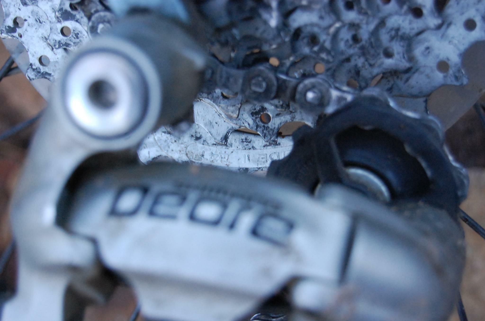 Gary Fisher Tassajara: Close-up image of a bicycle's rear derailleur and cassette, showing detailed components such as a metallic derailleur arm and the intricate gears of the cassette. The image captures the texture and wear of the parts, indicating usage. The background is slightly blurred, emphasizing the mechanical elements in focus.