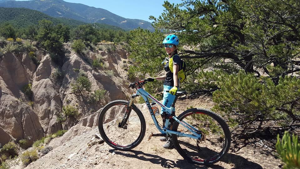 Trek Fuel EX 8: A person in a blue helmet and sunglasses stands next to a mountain bike on a rocky trail, surrounded by green trees and rugged terrain. In the background, mountains rise under a clear blue sky. The individual appears prepared for an outdoor biking adventure.