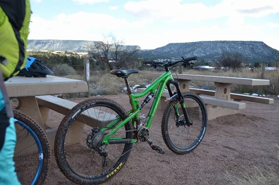 Santa Cruz Bronson: A green mountain bike is parked next to a picnic table in a sandy outdoor area, with scenic mountains in the background under a partly cloudy sky. The bike shows signs of recent use, with muddy tires. A blue helmet is visible on the table nearby.