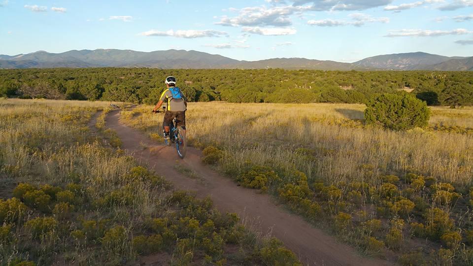 A person riding a mountain bike along a dirt trail through a grassy landscape, surrounded by shrubbery and mountains in the distance under a partly cloudy sky. La Tierra mountain bike trail.