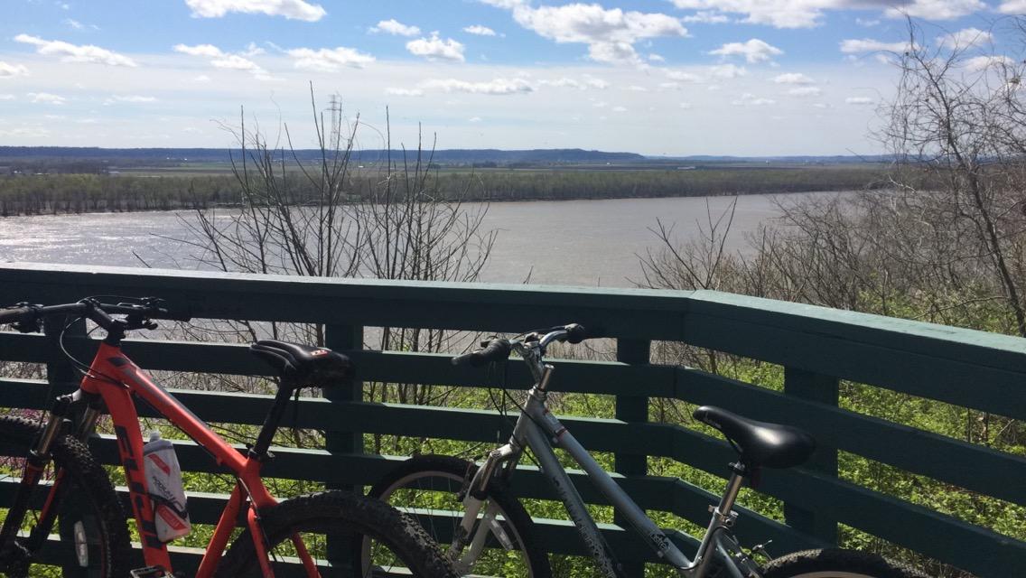 Two bicycles are resting against a wooden railing that overlooks a wide river. The scene is set under a bright blue sky with scattered clouds, showcasing a peaceful landscape of greenery along the riverbank and distant hills in the background. Bee Tree Park mountain bike trail.