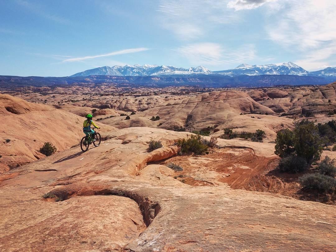 A mountain biker in bright green gear rides across a rocky landscape with rolling hills and distant snow-capped mountains under a clear blue sky. The scene captures the rugged beauty of the outdoors, highlighting the adventurous spirit of biking in nature. Slickrock mountain bike trail.