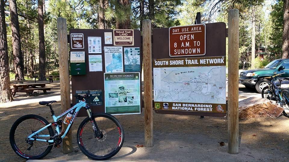 Trek Fuel EX 8: A mountain bike rests against a wooden information board in a forested area. The board displays various signs, including a caution sign about snakes, a welcome message, trail maps for the South Shore Trail Network, and visitor guidelines. Surrounding the board are picnic tables and tall pine trees, with a vehicle parked nearby. A sign indicates that the area is open from 8 AM to sundown.