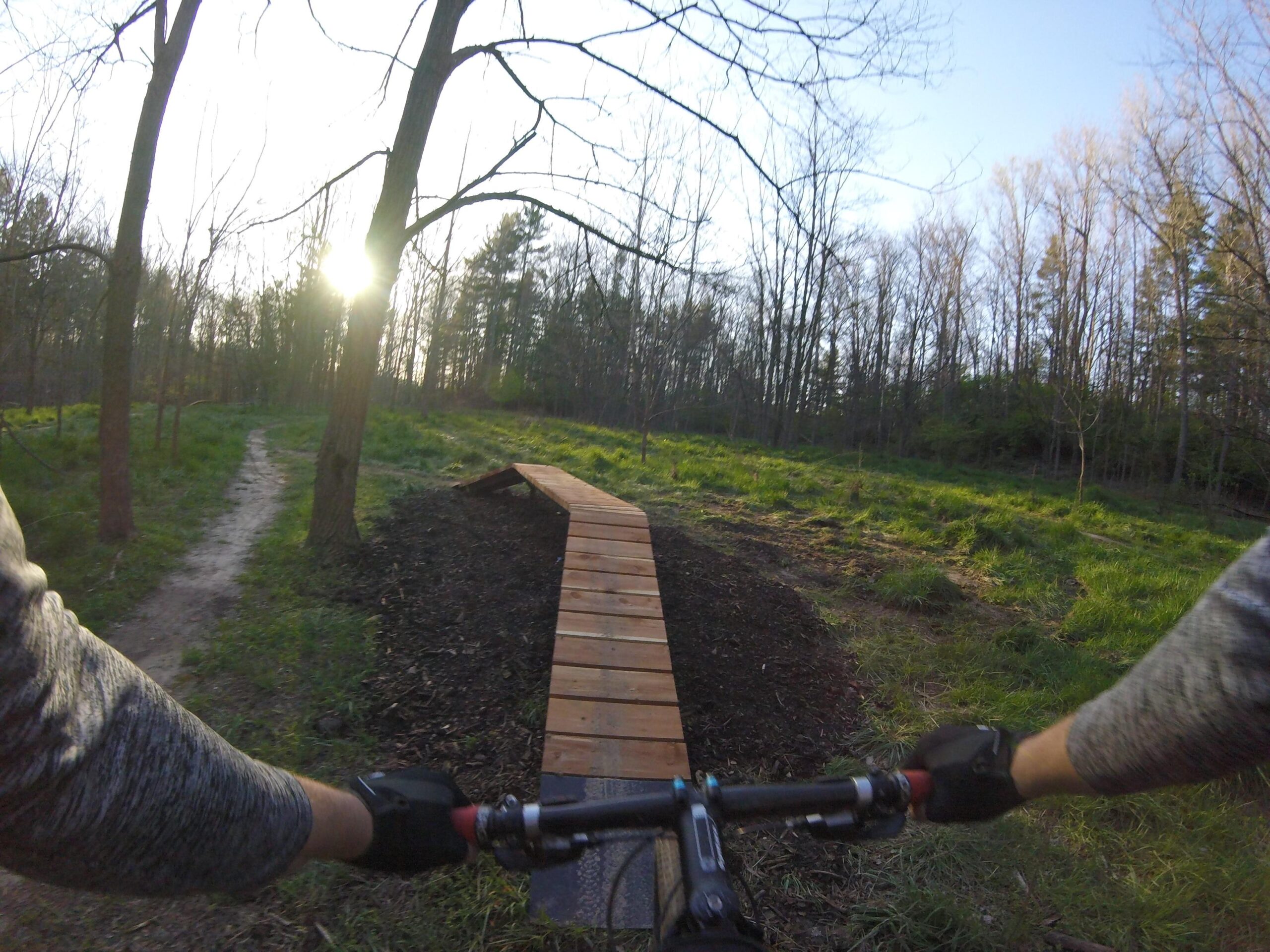 A view from the handlebars of a mountain bike, showing a wooden ramp and a winding dirt path through a forested area. Sunlight is peeking through the trees, illuminating the green grass and vibrant foliage in the background. Griffin Bike Park mountain bike trail.