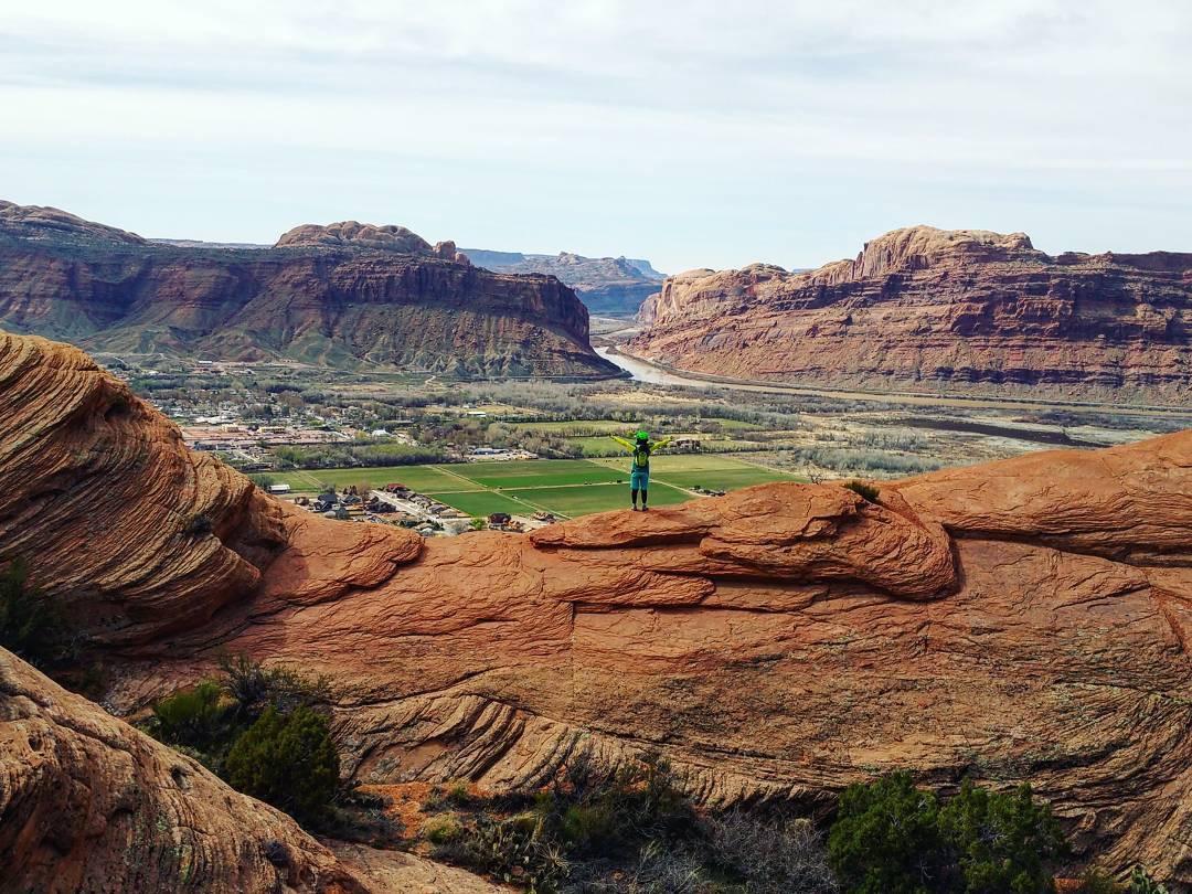 A person stands on a rocky outcrop overlooking a vast landscape of red rock formations, green fields, and a winding river below. The scene captures the natural beauty of the area, with rolling hills in the background and a cloudy sky above. Slickrock mountain bike trail.