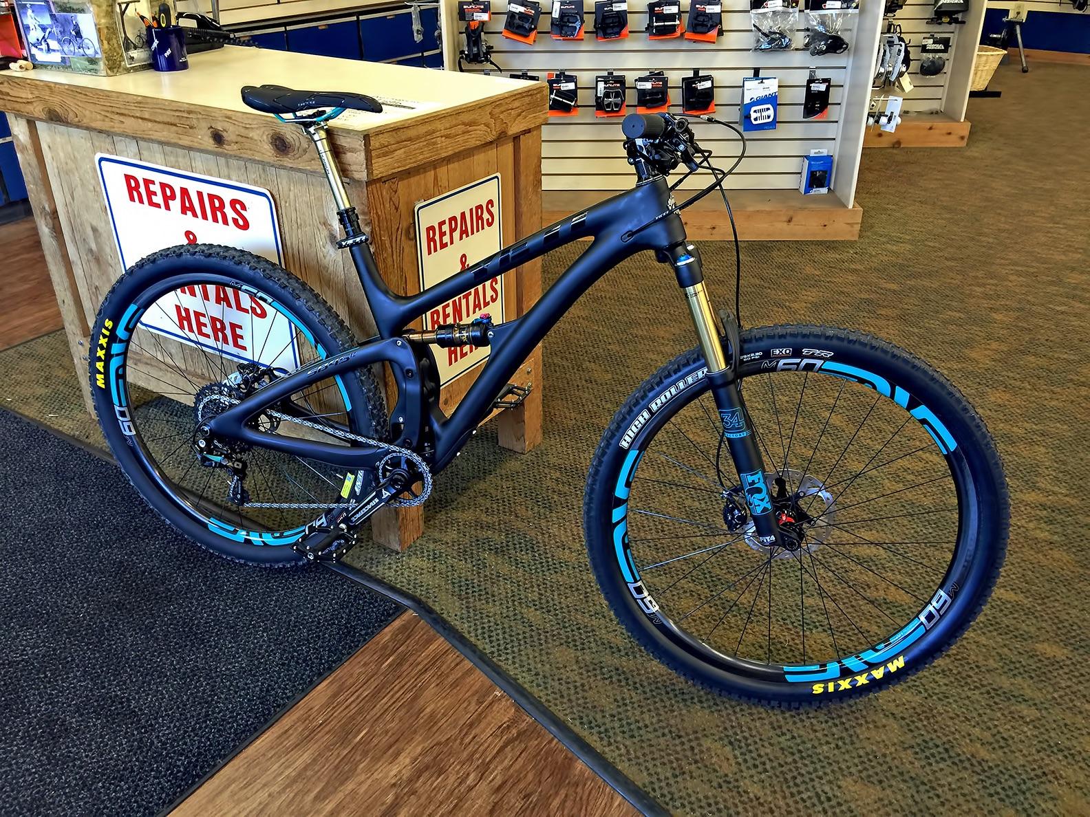 Yeti SB4.5c: A high-quality mountain bike displayed inside a rental shop. The bike features a lightweight black frame with blue accents, thick tires labeled "Maxxis," and front suspension. In the background, a wooden counter showcases signs for repairs and rentals. Various biking accessories and equipment are arranged on the wall behind the counter.