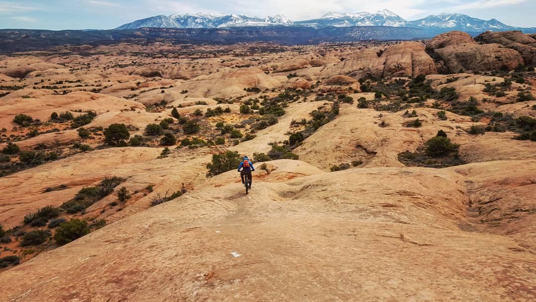 A mountain biker descends a rocky, arid landscape with scattered vegetation, set against a backdrop of snow-capped mountains under a partly cloudy sky. Slickrock mountain bike trail.