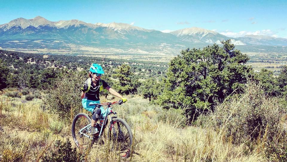 A mountain biker on a trail in a mountainous landscape, surrounded by greenery and shrubs. In the background, towering mountains rise under a clear blue sky. The biker is wearing a blue helmet and cycling gear, navigating the terrain on a mountain bike. Methodist Mountain mountain bike trail.